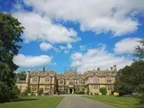 Large stone manor house with a long driveway under a bright blue sky with puffy white clouds.
