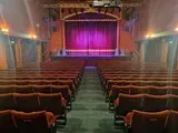 Interior of a theater. Rows of red seats face a stage with a purple curtain, under a gold arch.