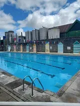 Outdoor swimming pool with blue water and lane markers. Colorful changing sheds line the edge, under a cloudy sky.
