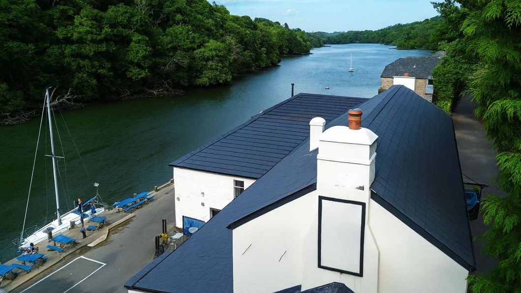 White building with dark blue roof, next to water and trees. Sailboat docked.