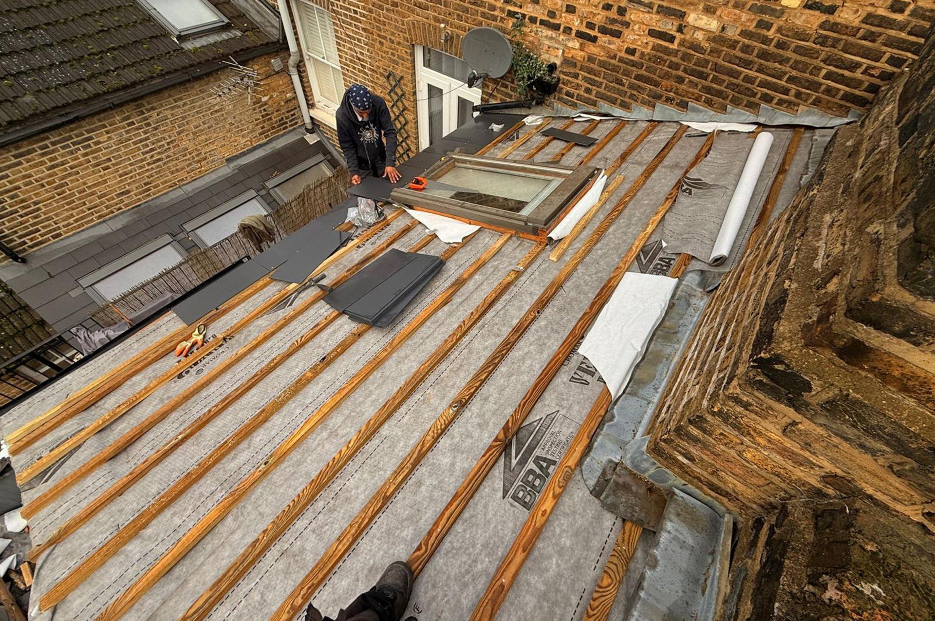 Roofer repairing a partially dismantled flat roof with exposed wooden beams, a skylight, and adjacent buildings.
