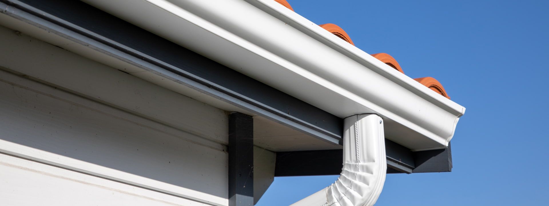 White rain gutter on a house with dark supports, terracotta tiles, and a blue sky.