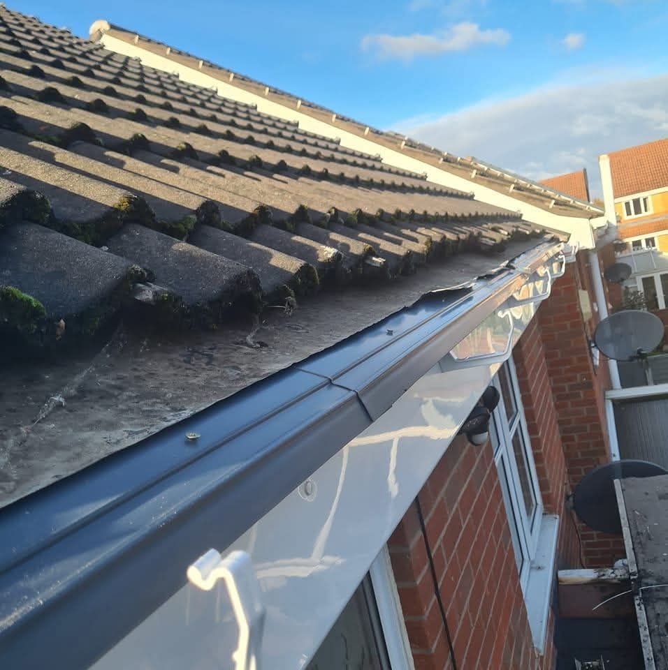 A close-up of a house roof edge with dark brown roof tiles and a new, dark grey guttering system installed above a window.