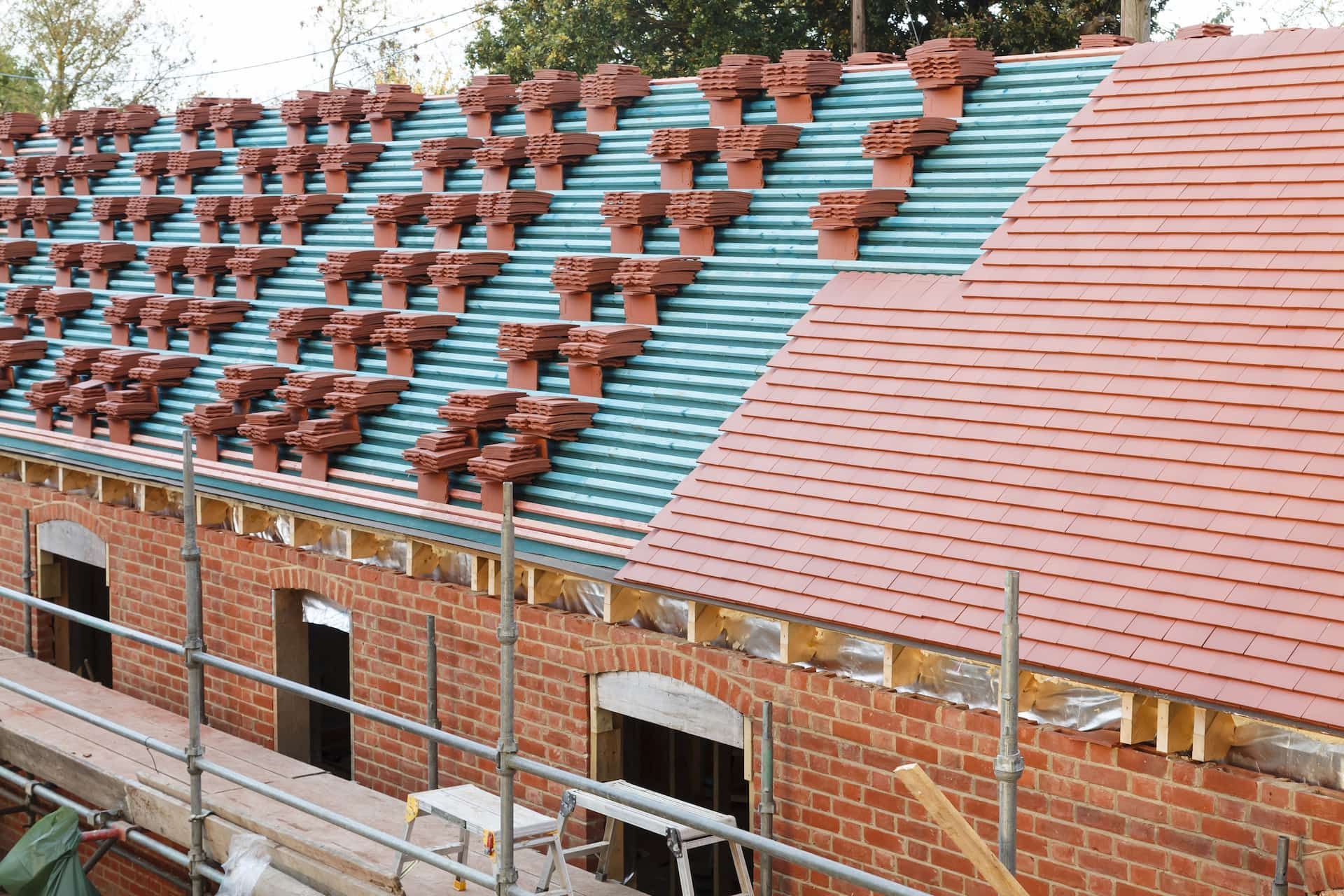 Brick building under construction, showing red tiles being installed on a roof with visible scaffolding.