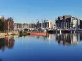 Waterfront view with calm water reflecting buildings, boats, and a clear blue sky.