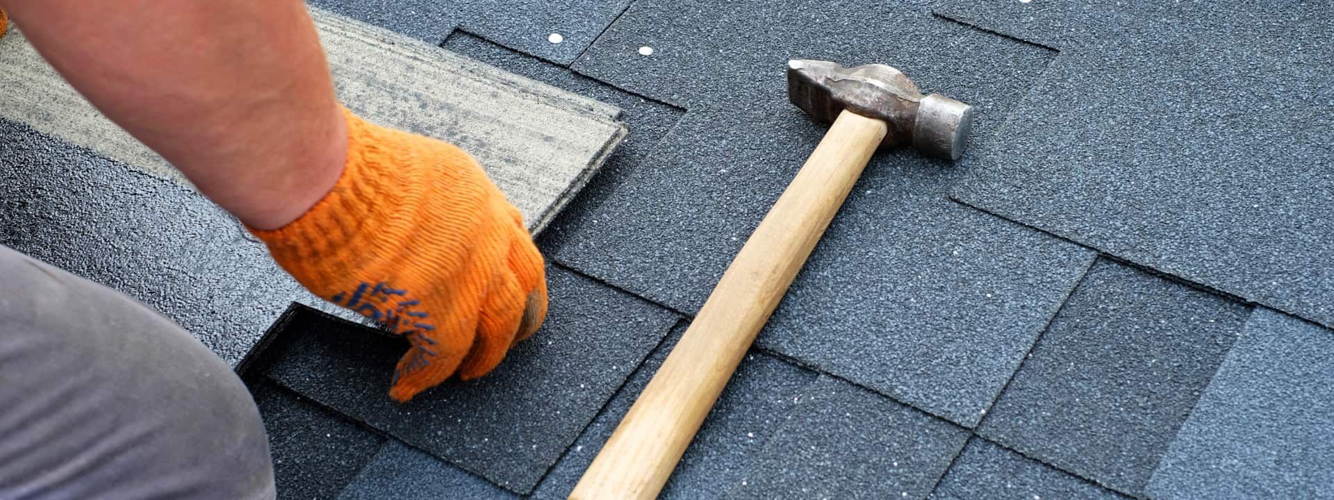A person with an orange-gloved hand placing a shingle. A hammer rests on a blue shingled roof.