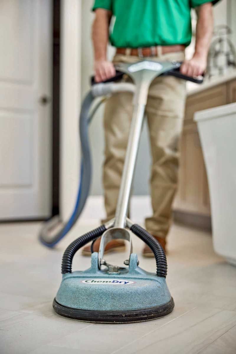 Person in green shirt and khaki pants using a floor cleaning machine in a bathroom.