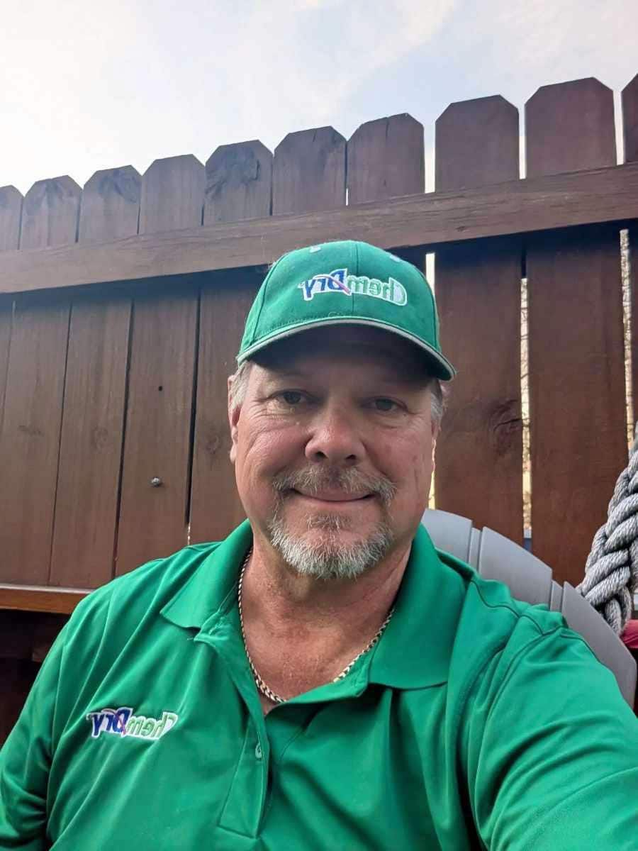 A Chem-Dry technician wearing a green hat and shirt, smiling, in front of a wooden fence.