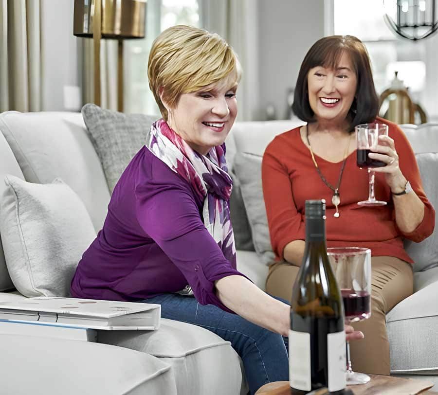 Two women on a couch, one pouring wine, looking at a photo album in a brightly lit living room.