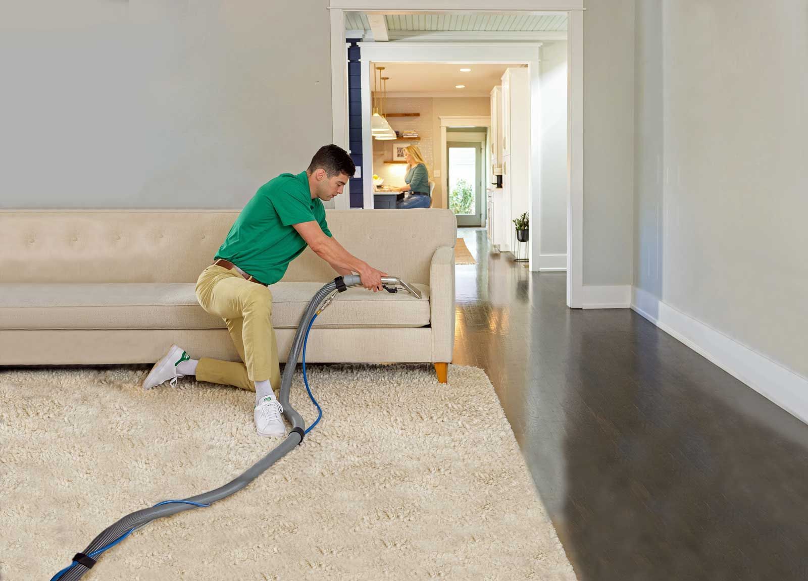 A Chem-Dry technician is cleaning a beige sofa with a hose on a light-colored rug in a home.