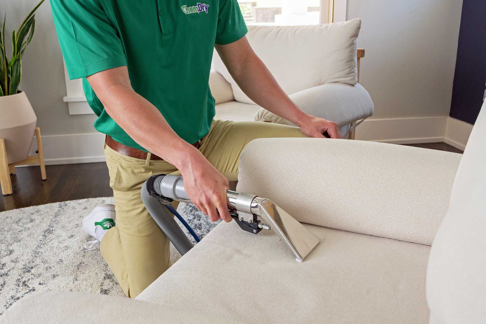 A Chem-Dry technician in a green shirt is cleaning a white sofa with a vacuum in a living room.