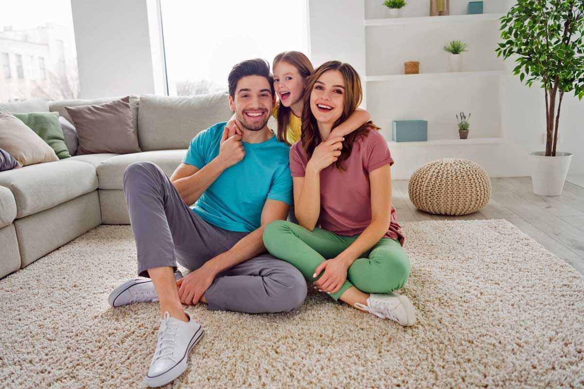 Family of three smiling, sitting on a fluffy rug in a bright living room; the little girl has her arms around her mom's neck.