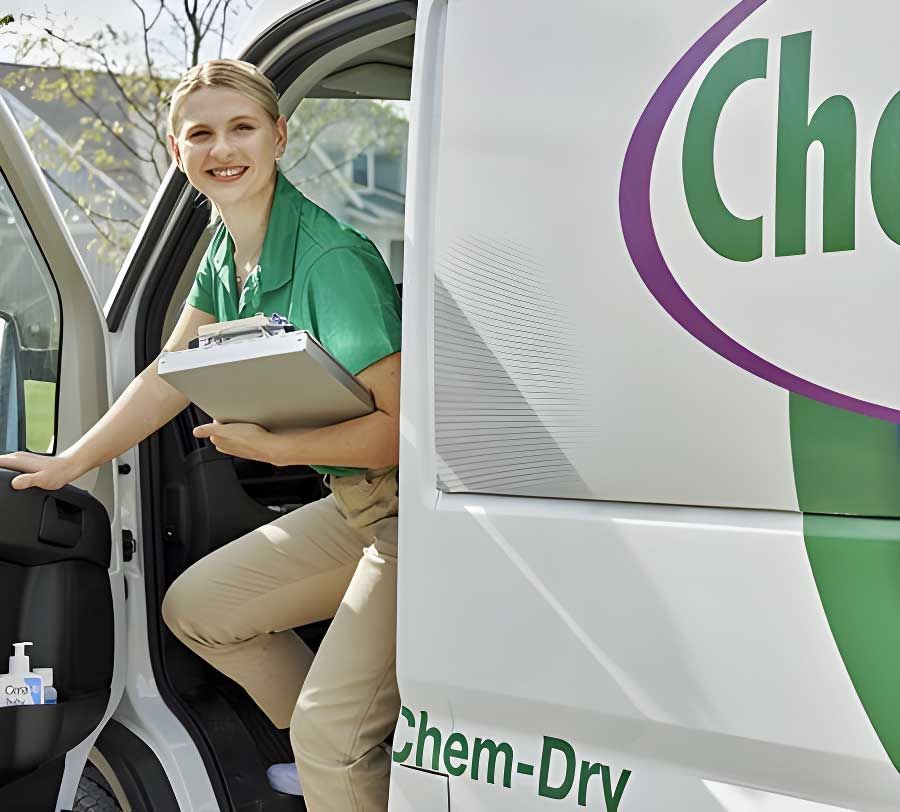 A Chem-Dry employee in a green shirt and khaki pants steps out of a Chem-Dry van, holding a clipboard and smiling.