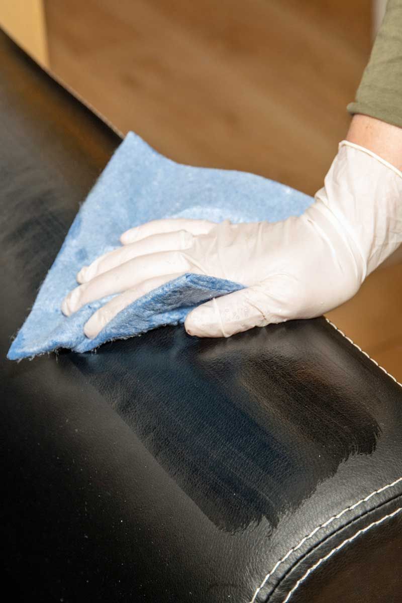 Person wearing white gloves wiping down black leather furniture with a blue cloth.
