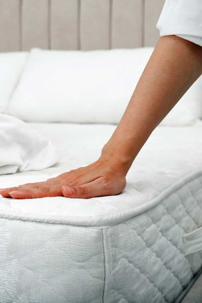 A hand presses down on a white mattress in a bedroom setting. The mattress is covered with white bedding.