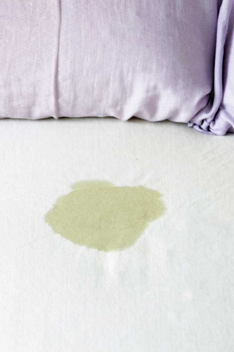 A bed with a white sheet and a light purple pillow. A wet, yellow stain is on the sheet.