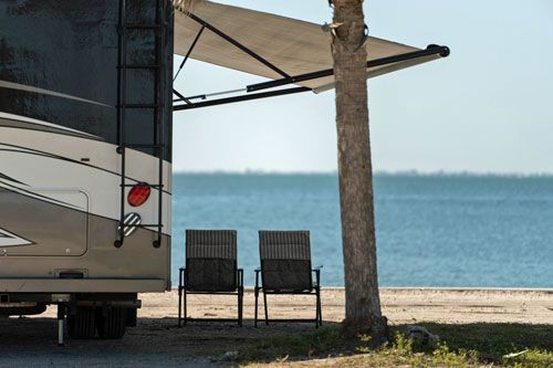 RV parked near beach with awning extended; two chairs sit under palm tree overlooking water.