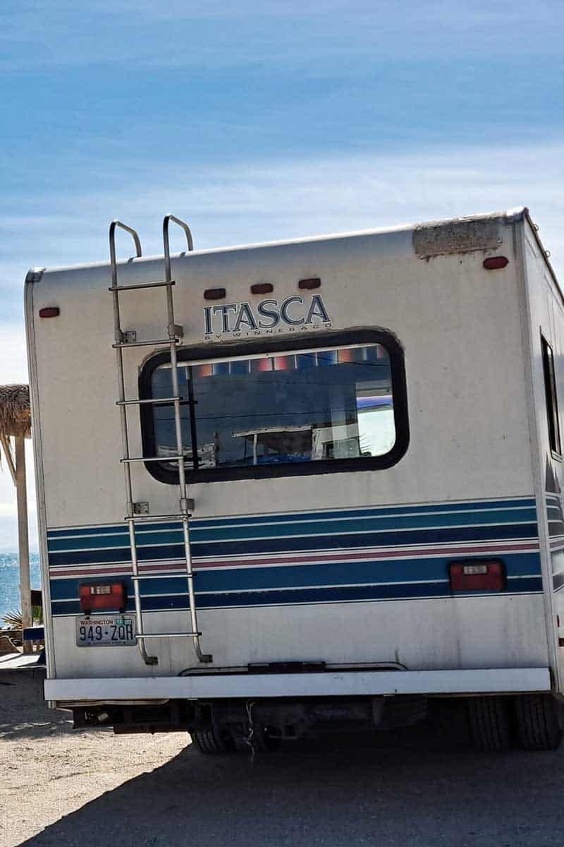 Back of a weathered Itasca RV with ladder, parked outdoors near a beach; the sky is blue.