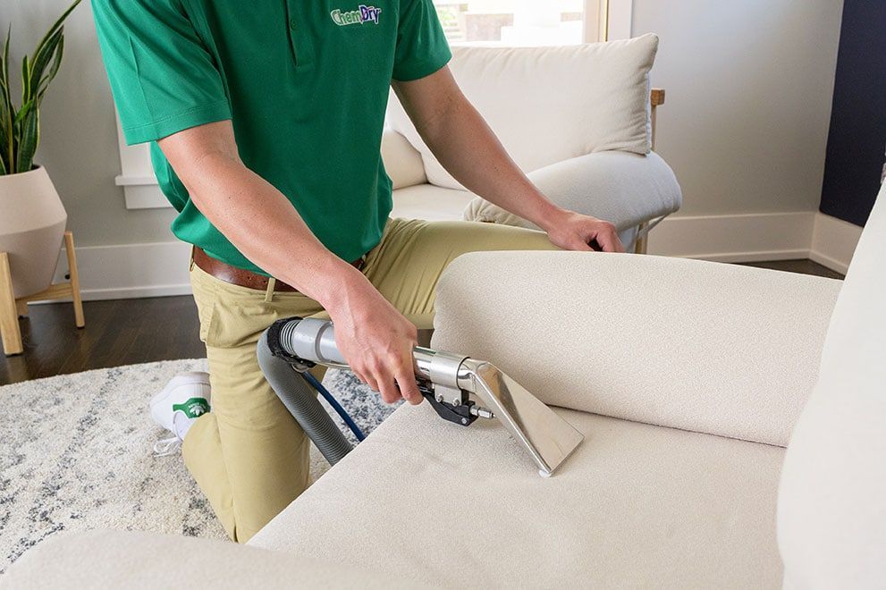 A Chem-Dry technician in a green shirt and khaki pants cleans a white couch with an upholstery cleaner.