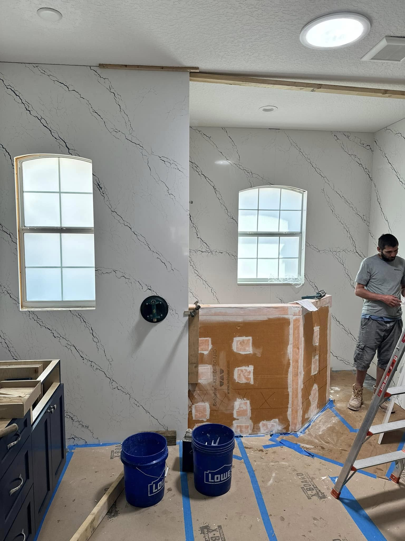 Kitchen under renovation: White walls with dark streaks, two windows, blue buckets, man on ladder.