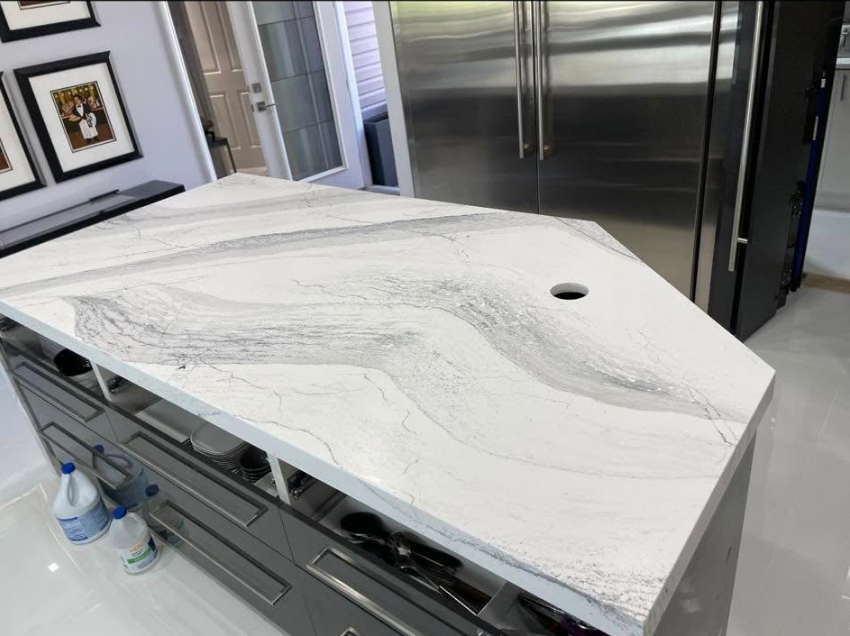 Kitchen island with white and gray marbled countertop and gray cabinets. A stainless steel refrigerator is visible.