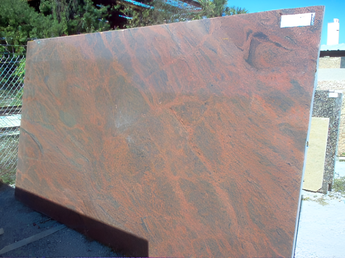 Large slab of red granite with dark veining, leaning against a chain-link fence outdoors.