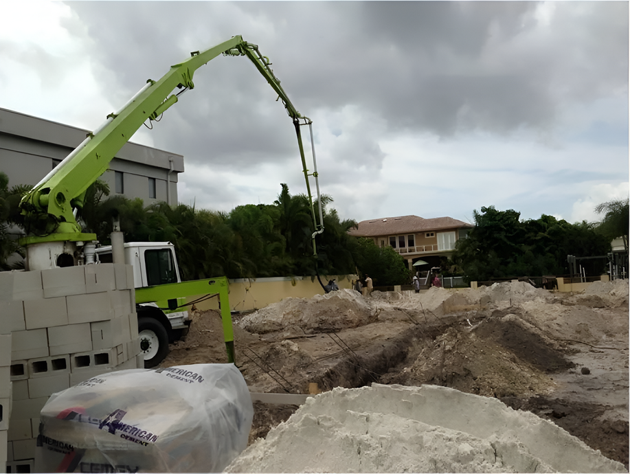 Concrete pump truck pouring concrete at a construction site near a house, with cloudy sky.