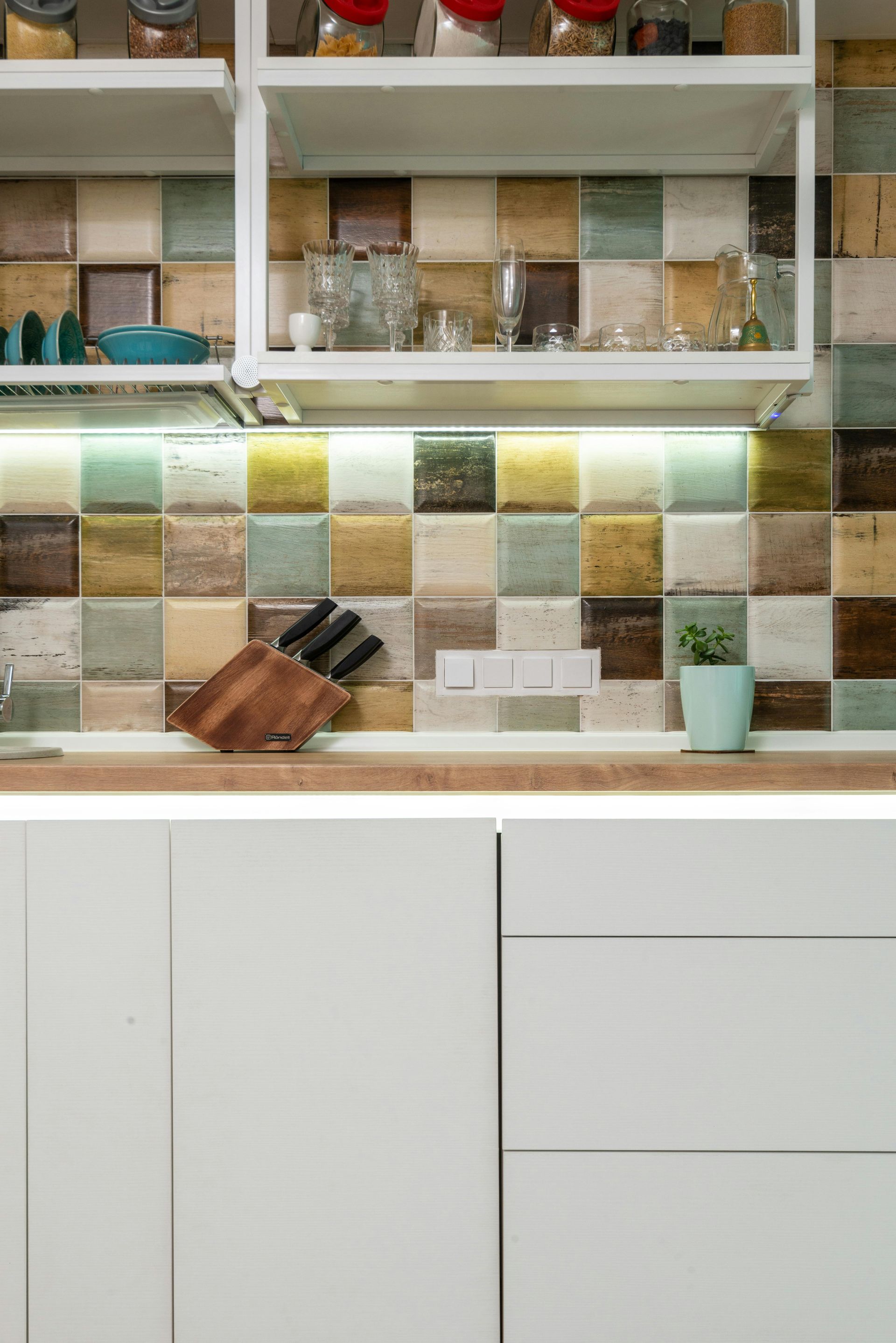 Kitchen backsplash with colorful square tiles, white cabinets, wooden countertop and knife block.