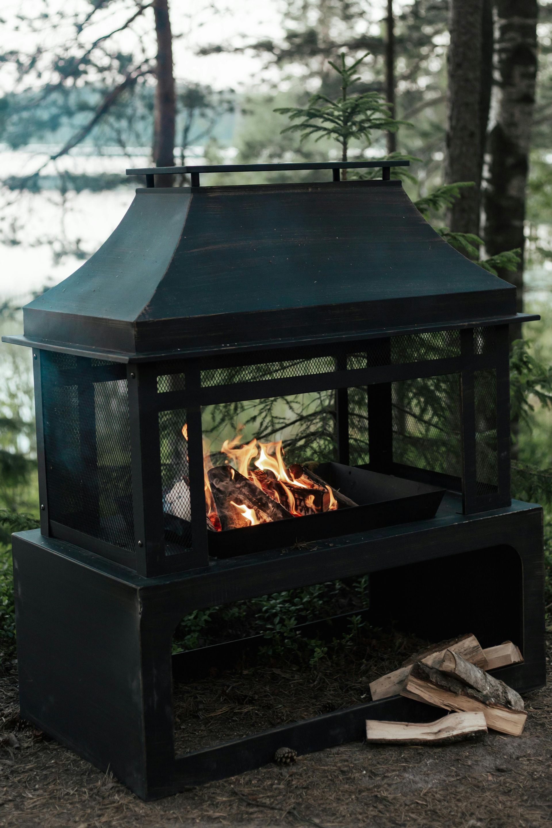 Black metal outdoor fireplace with burning wood, next to a pile of logs, in a forest setting.