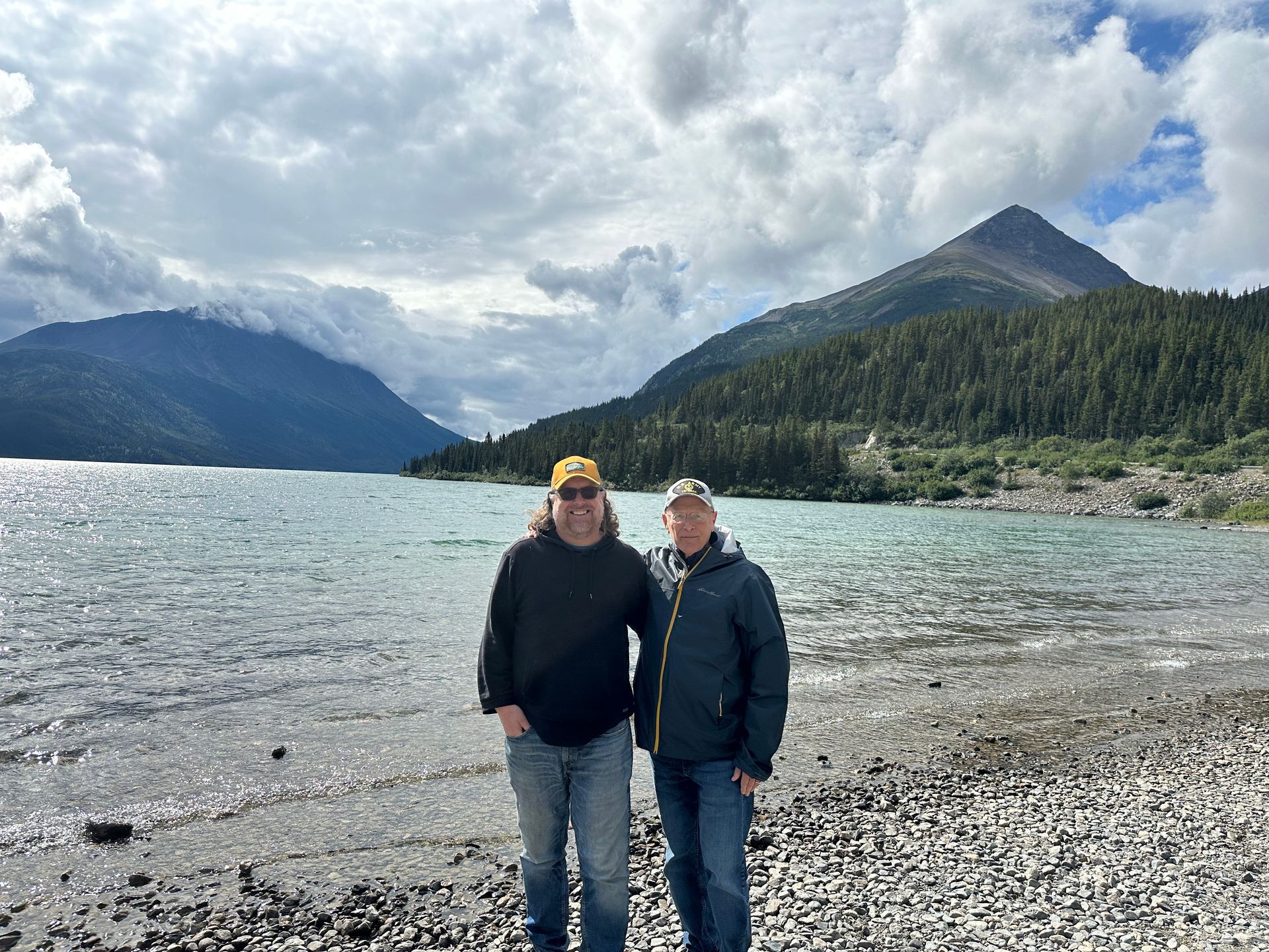 Two men are standing on a rocky beach next to a lake.