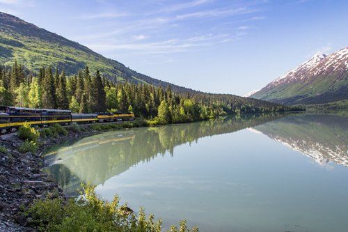 A train is going down the tracks next to a lake with mountains in the background.