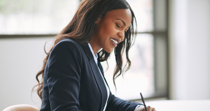 A woman in a suit is sitting at a desk writing on a piece of paper.