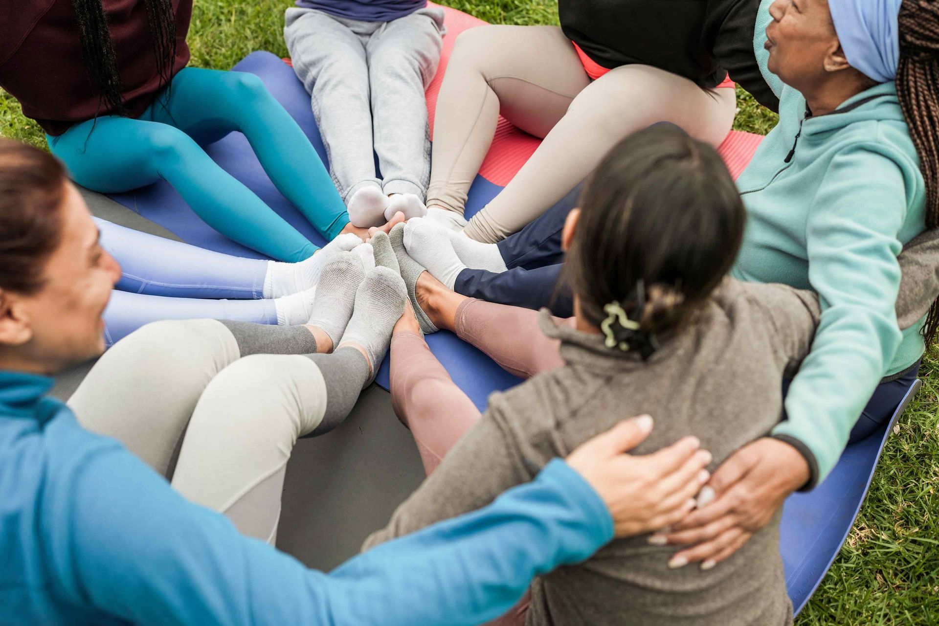 Group of people sitting in a circle, legs touching, on a mat in a grassy area.