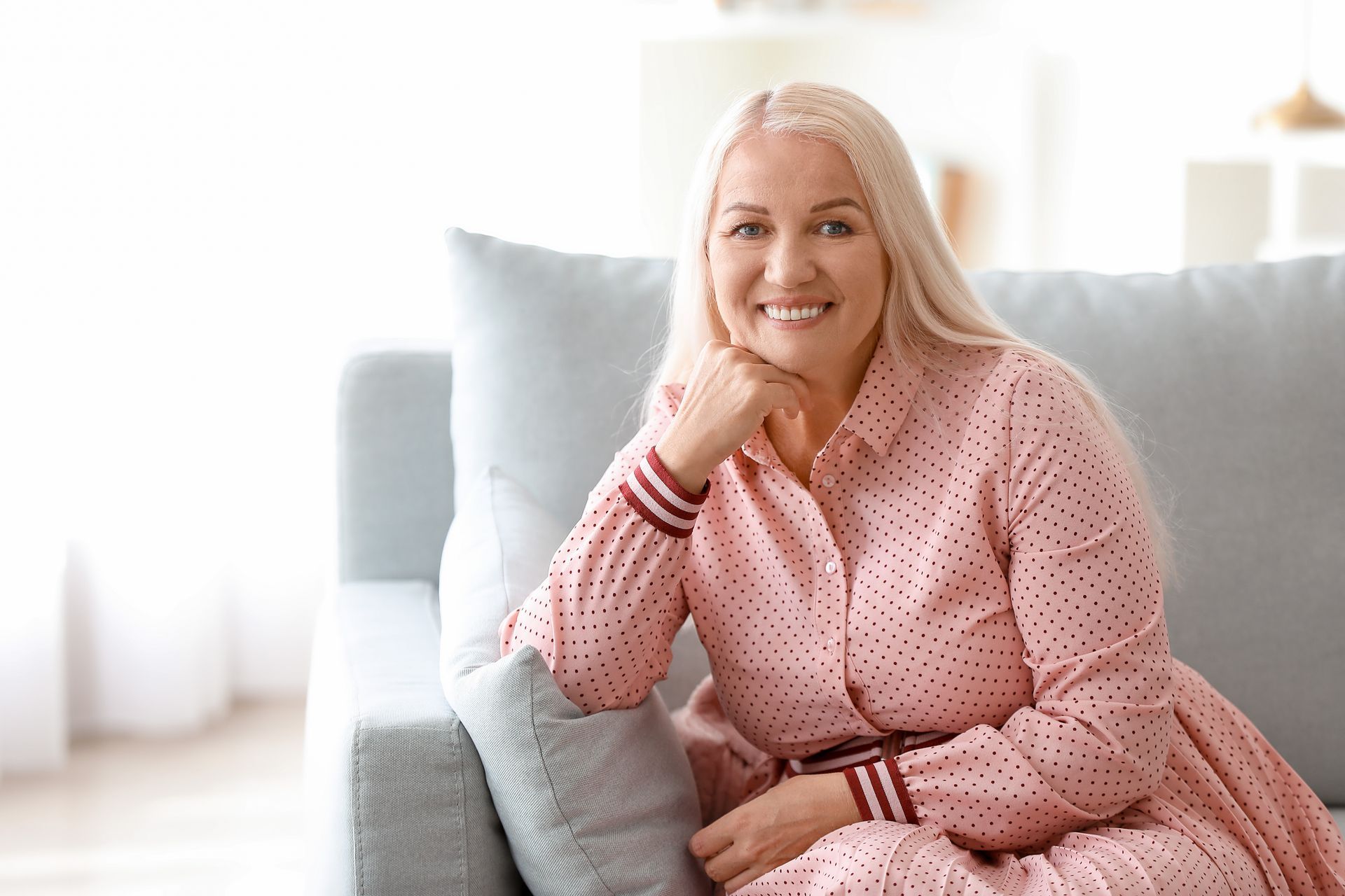 Woman with long blonde hair, sitting on a light blue couch, wearing a pink dress, smiling, indoors. Woman with long blonde hair, sitting on a light blue couch, wearing a pink dress, smiling, indoors.