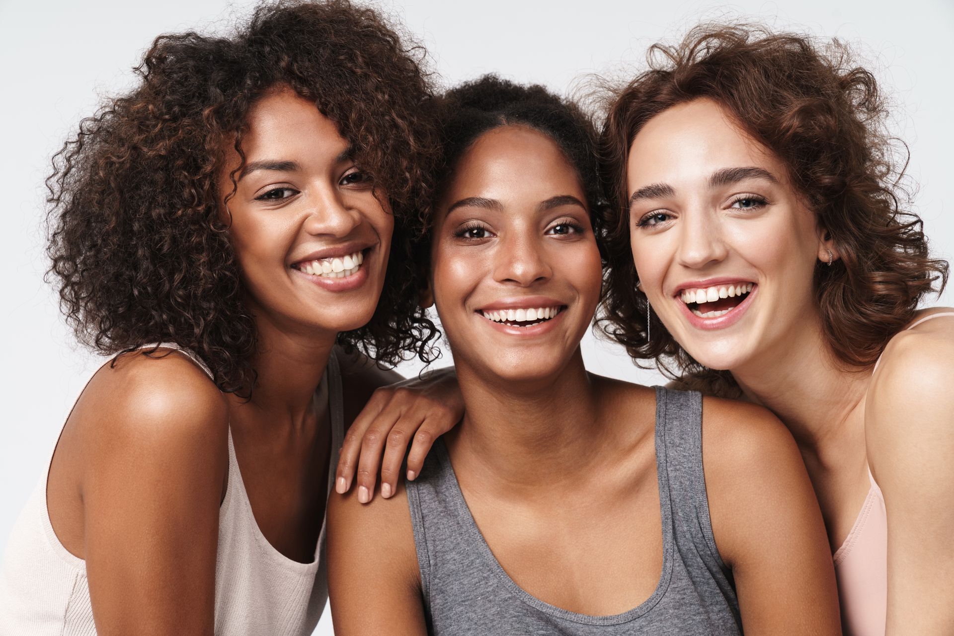 Three smiling women with different skin tones, in tank tops, posing together.