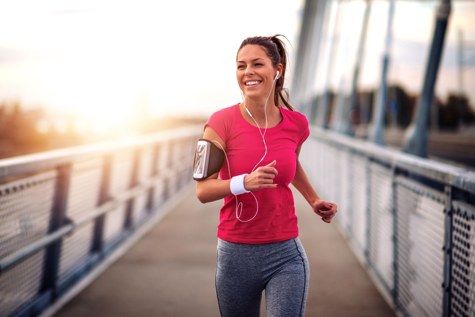 Woman running on a bridge, smiling, wearing red shirt, arm band, and earbuds. Sunlight in the background.