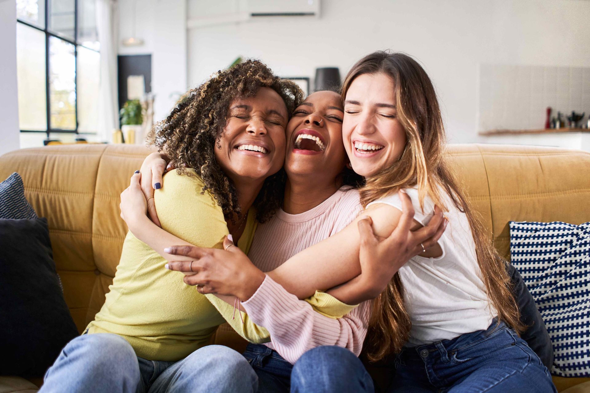 Three friends laughing and hugging on a yellow couch in a living room.