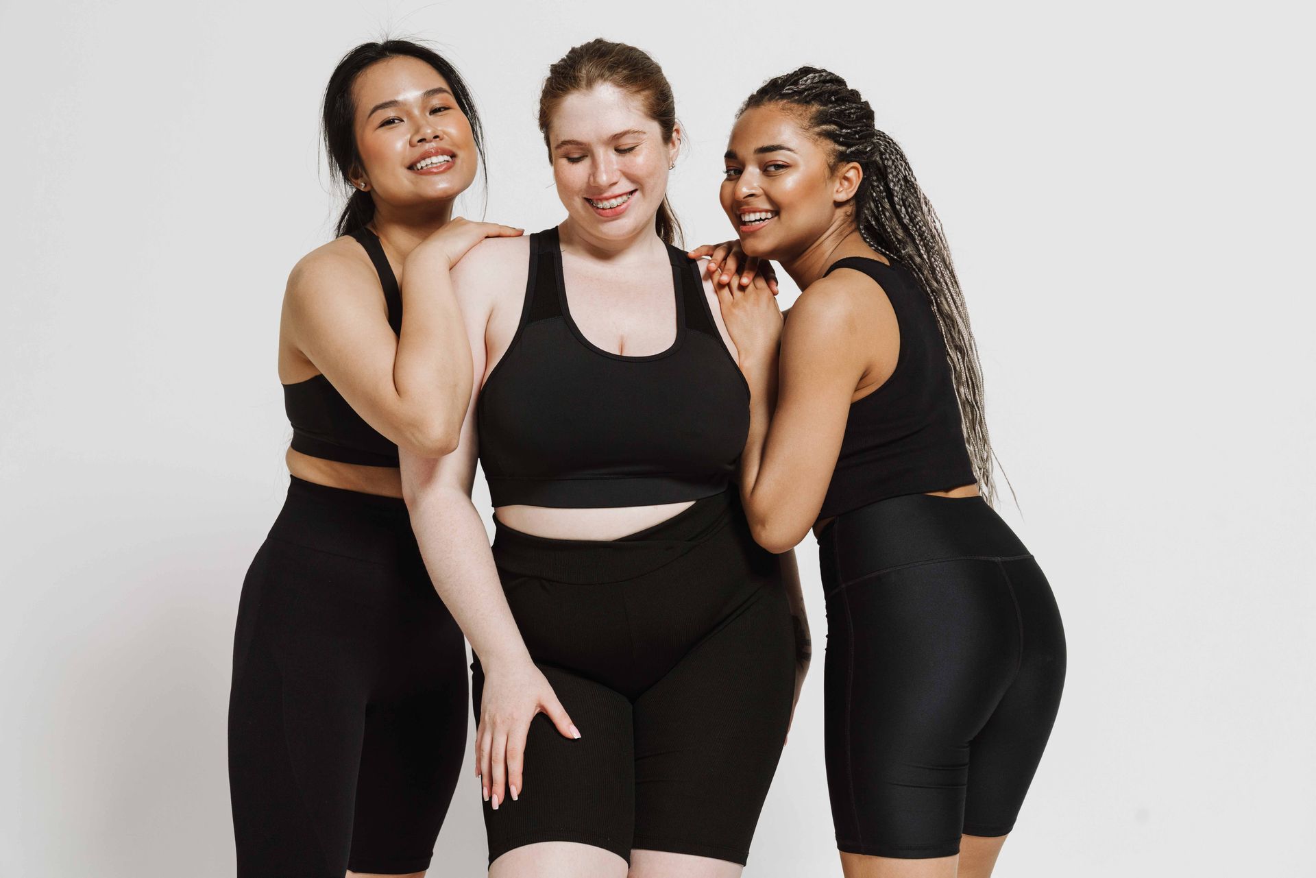 Three women in black athletic wear, smiling and embracing against a white background. Three women in black athletic wear, smiling and embracing against a white background.
