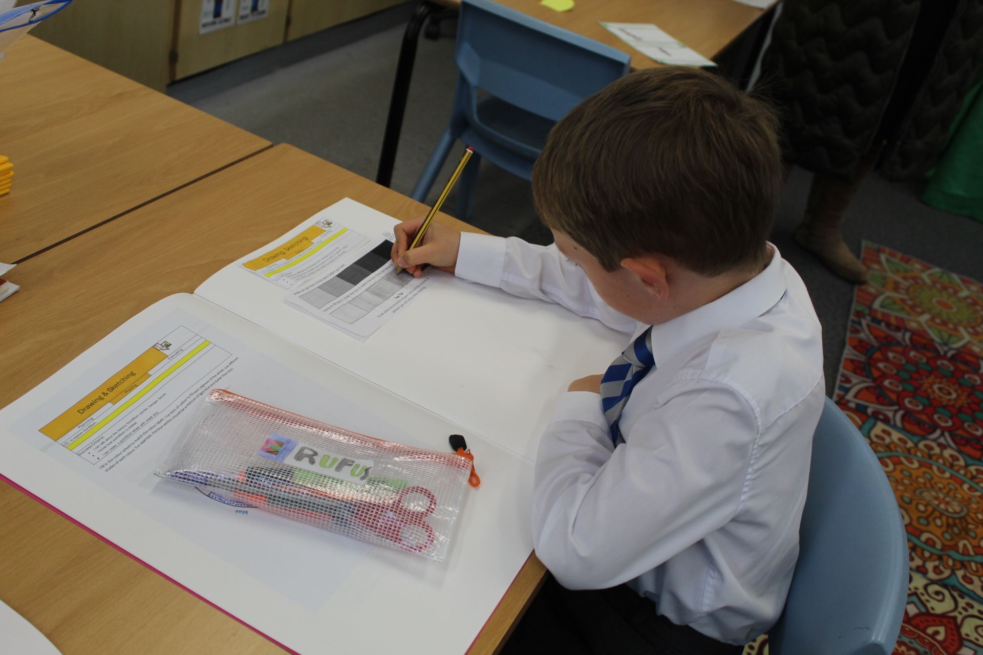 Two children in school uniforms seated at desks. Looking toward camera, holding folders.
