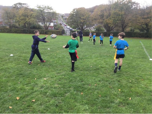 Children playing tag rugby on a grassy field; one boy throwing the ball, others running, with trees in the background.