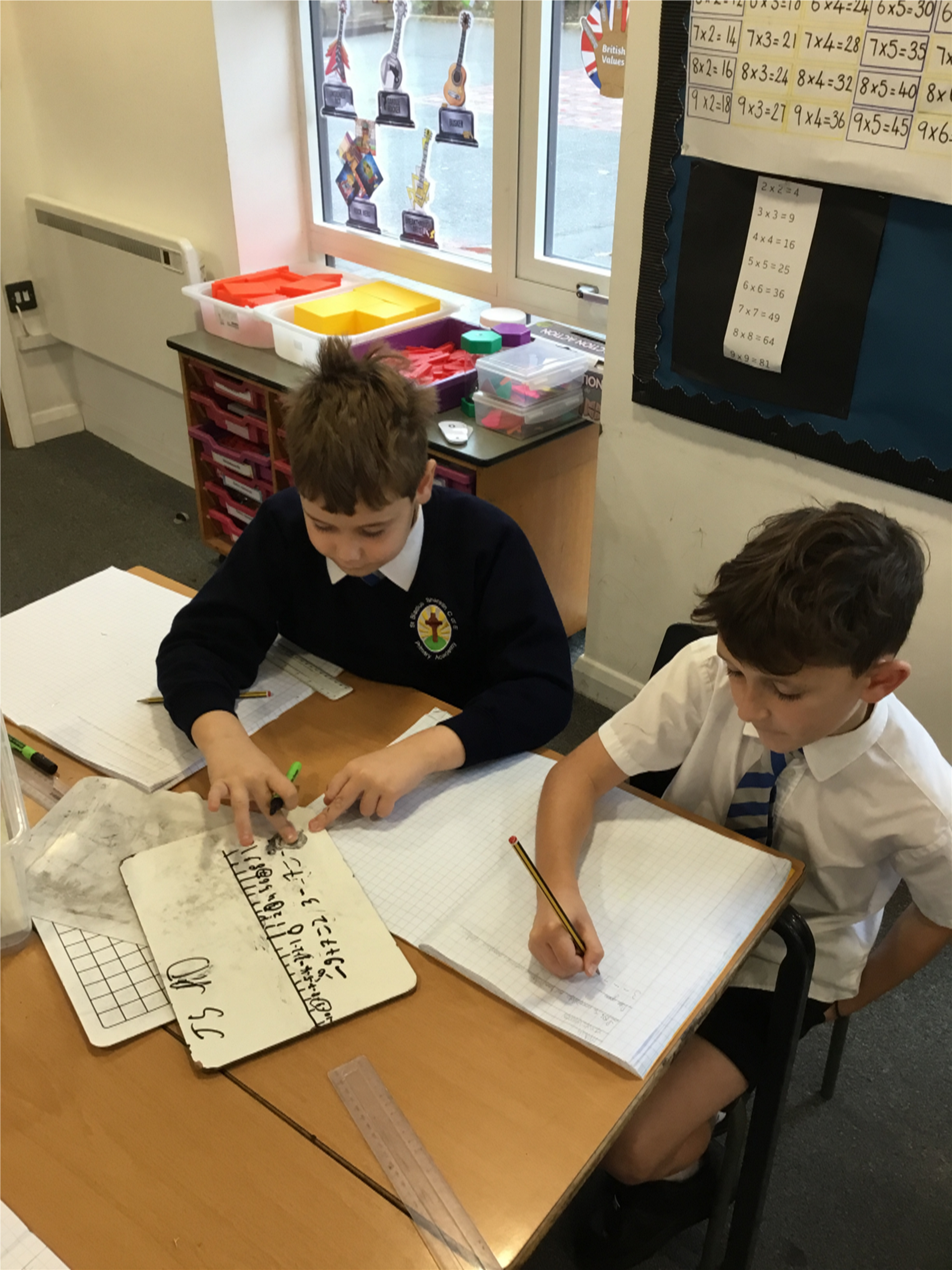 Two young boys are sitting at a desk in a classroom writing in notebooks.