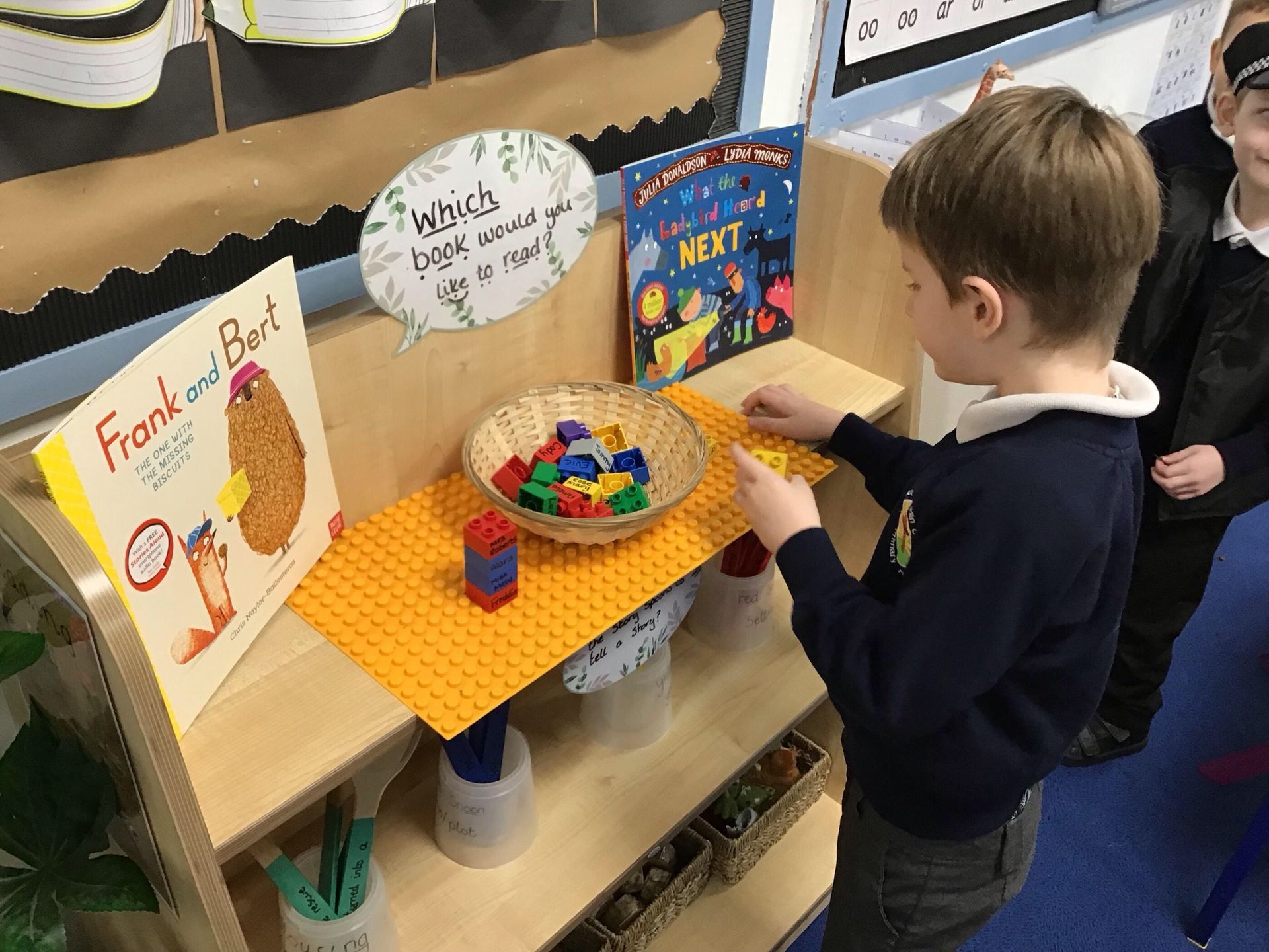 A child stands at a classroom shelf, building with small colorful blocks near a book titled 