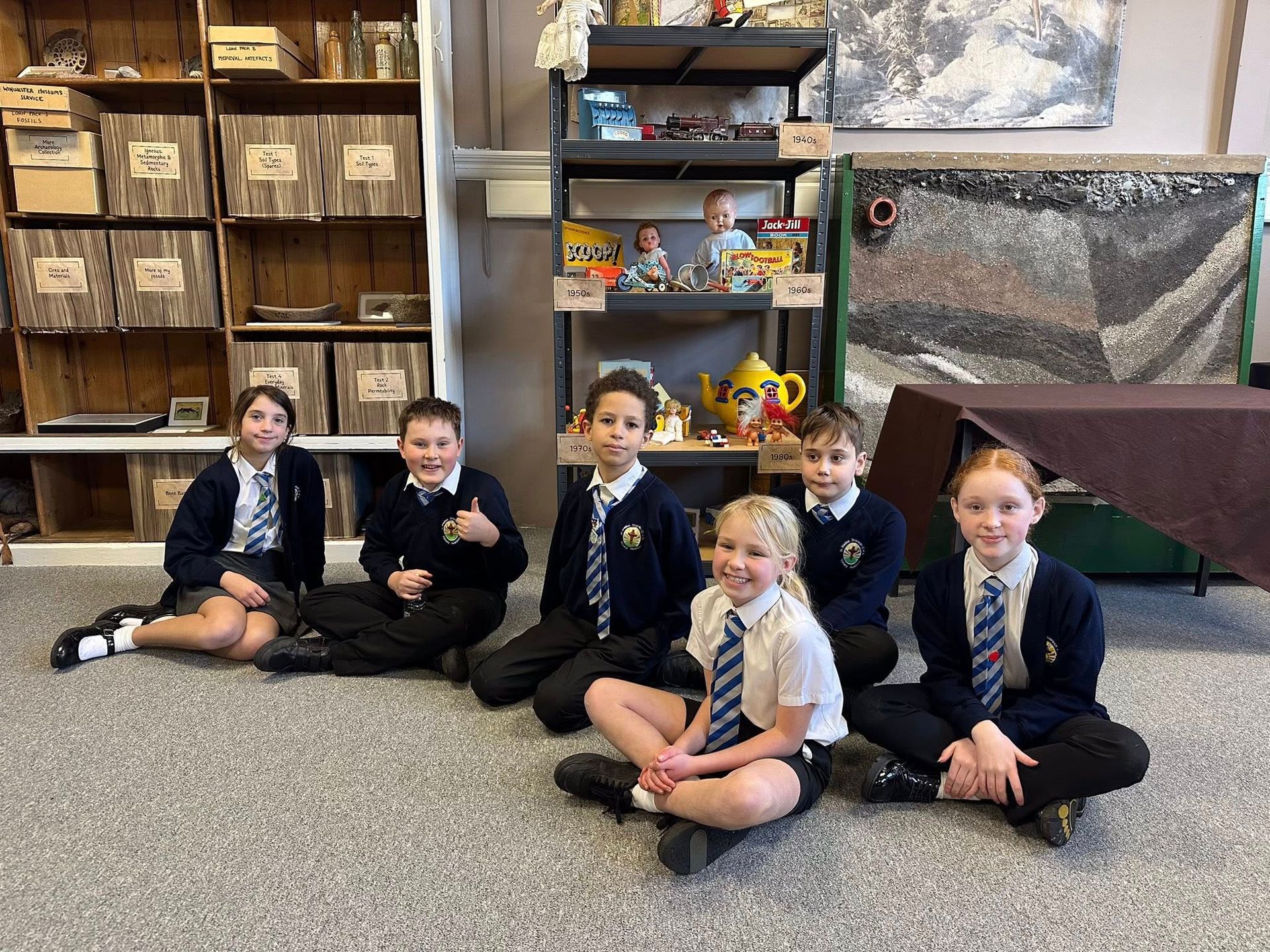 Six students in school uniforms sit on the floor in front of shelves filled with various objects.
