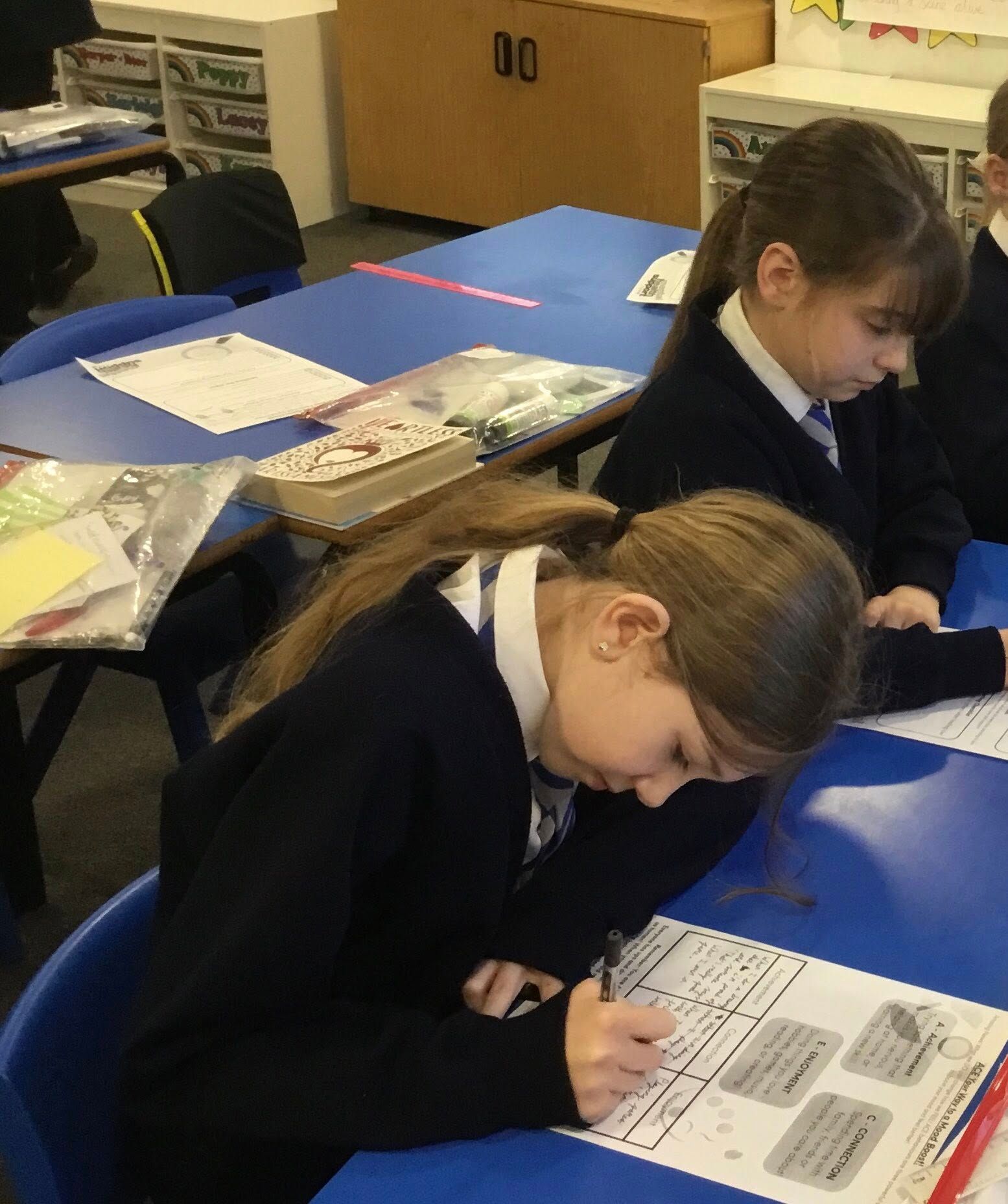 Two students in navy uniform writing at blue desks in a classroom.