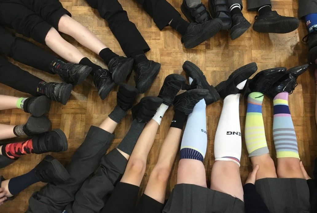 A circle of legs wearing black pants and shoes, some with colorful socks, on a wooden floor.