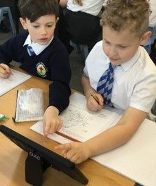 Two schoolchildren writing on worksheets at a desk, using a tablet in a classroom.