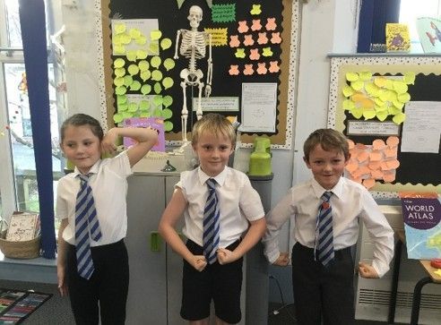Three children flexing their arms in a classroom with a skeleton model and posters on the wall.