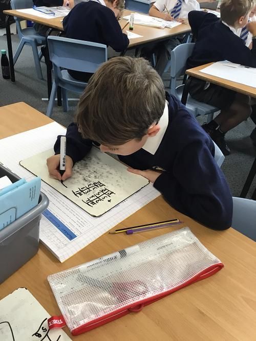 Boy in school uniform drawing in a notebook at a desk, with pencil case and other students in the background.