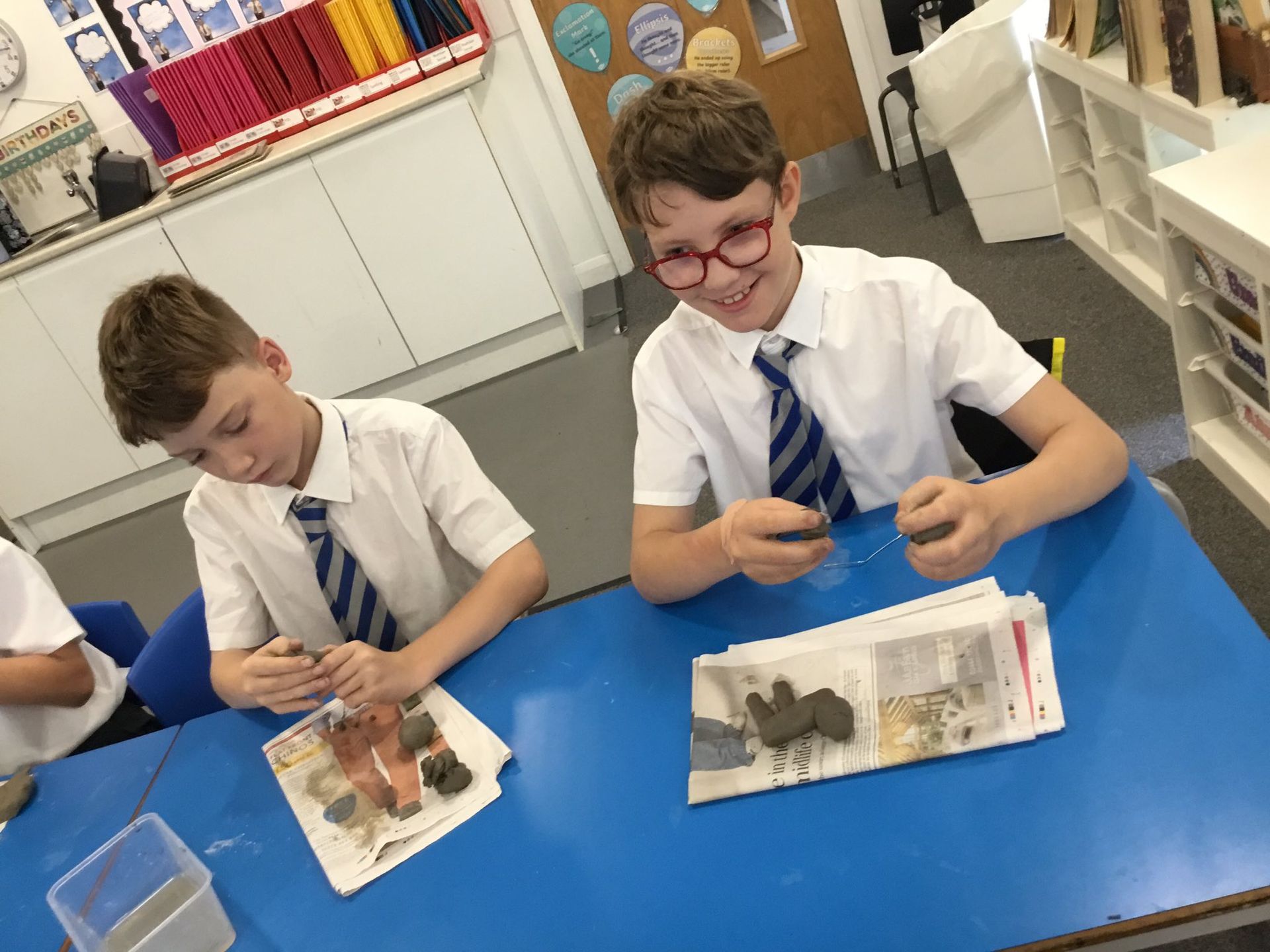 Two boys in school uniforms at a blue table, making something with clay. One smiles, wearing glasses.