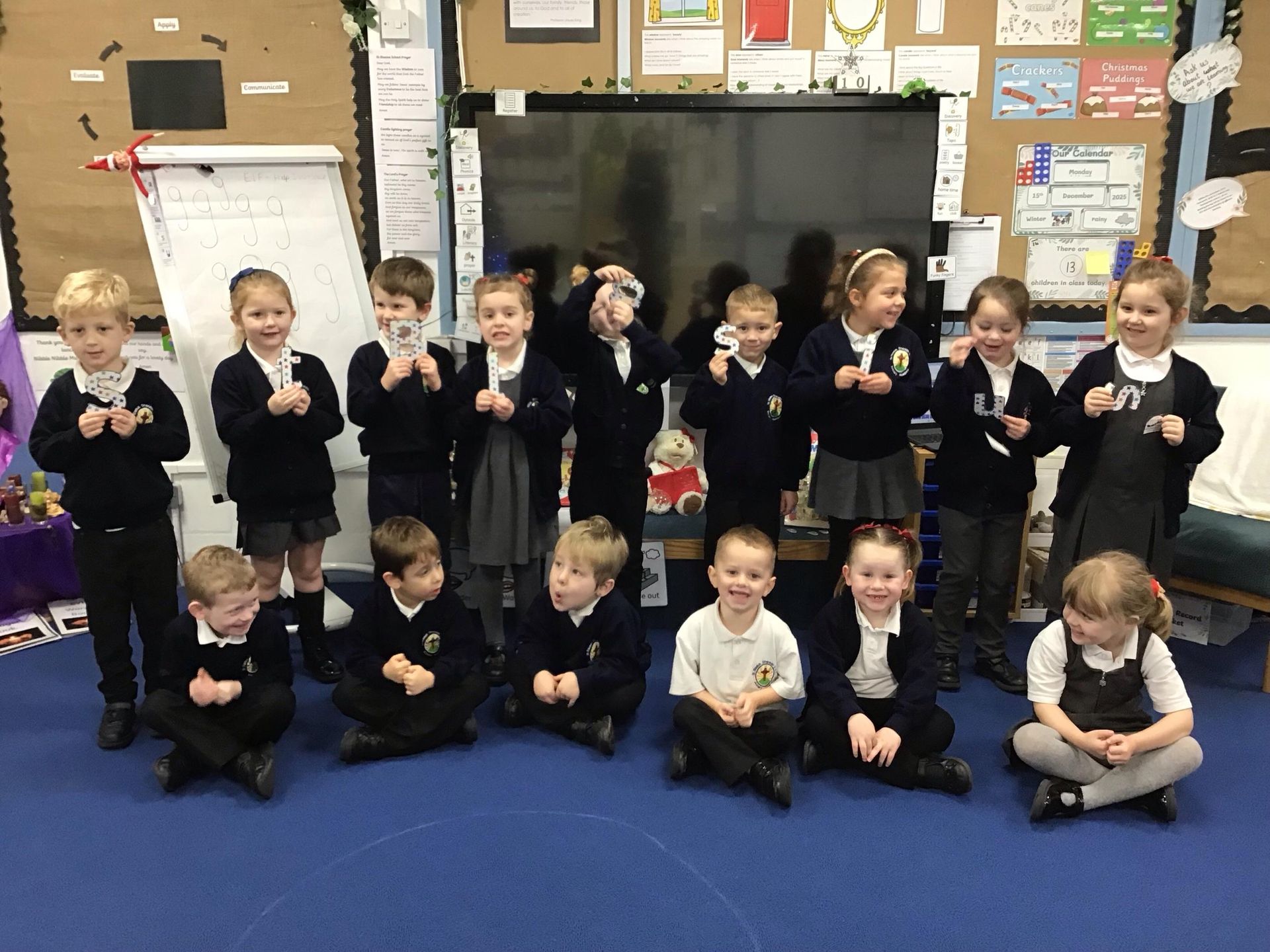 Children in a classroom, some sitting, all wearing uniforms, performing hand gestures. Background has posters and a whiteboard.